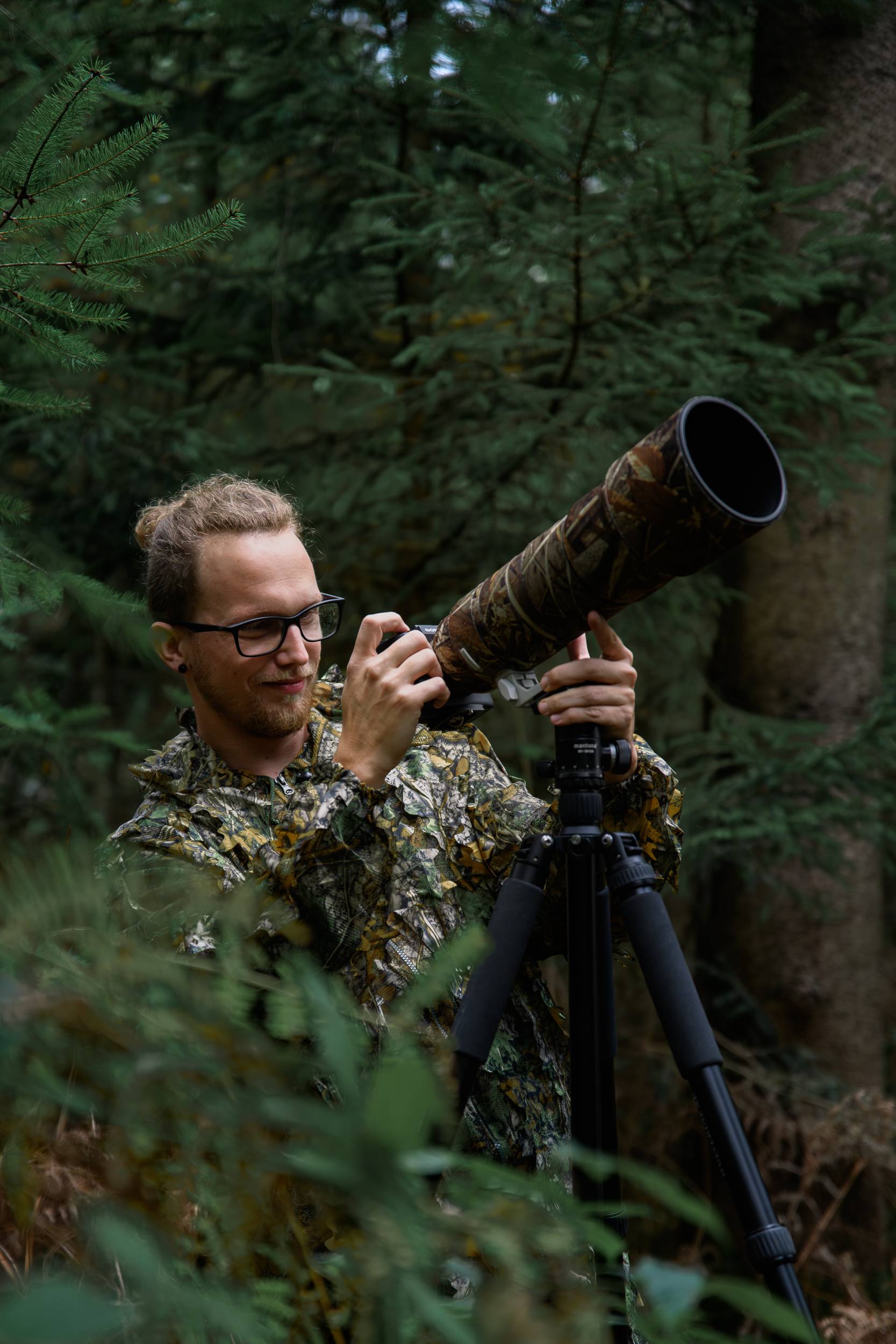 Rainer Lentjes, conservation and wildlife photographer, looking through a large telephoto camera lens on a tripod, surrounded by dense forest vegetation.