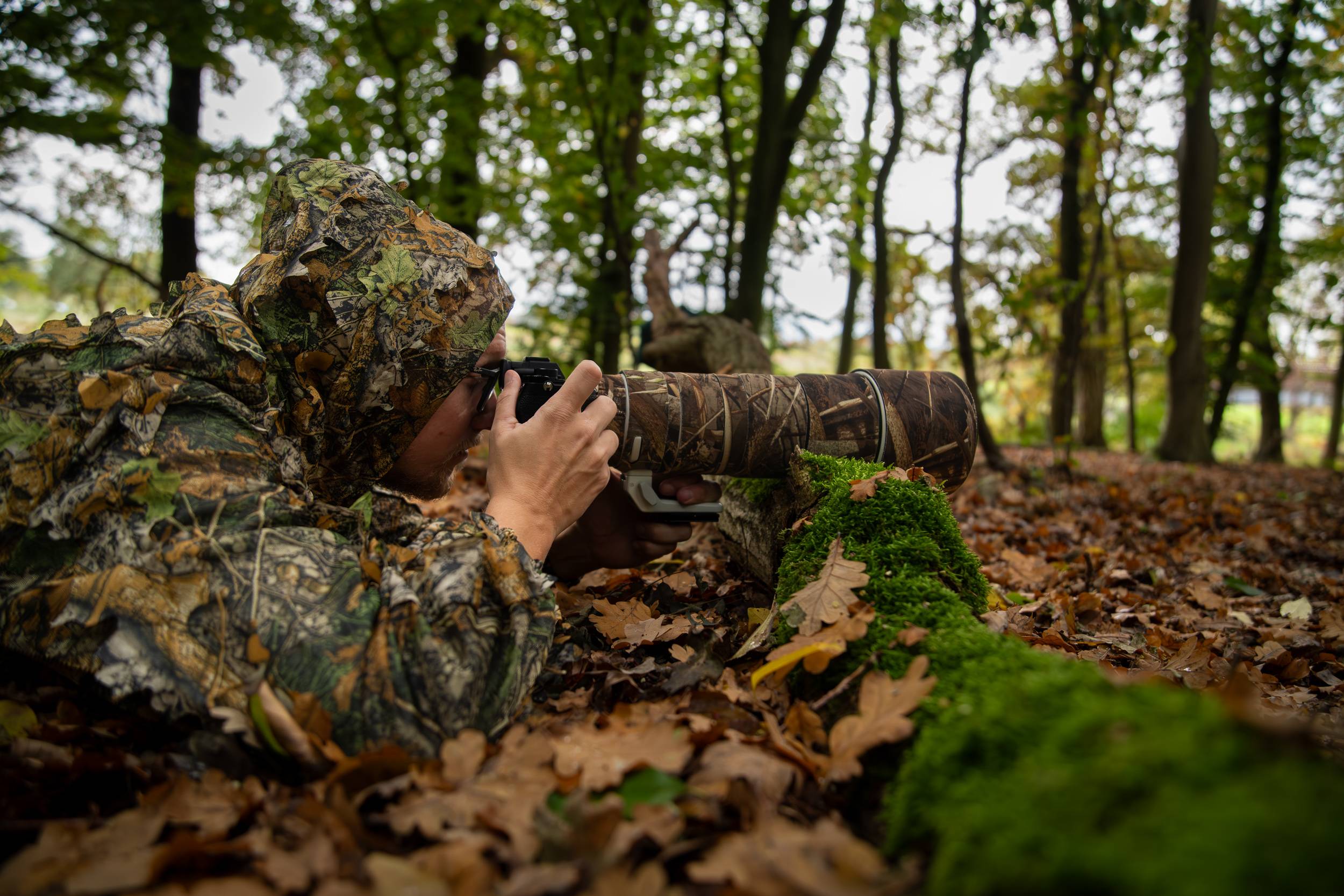 Rainer Lentjes wildlife photographer lying on the forest floor in camouflage gear capturing images through a telephoto lens.