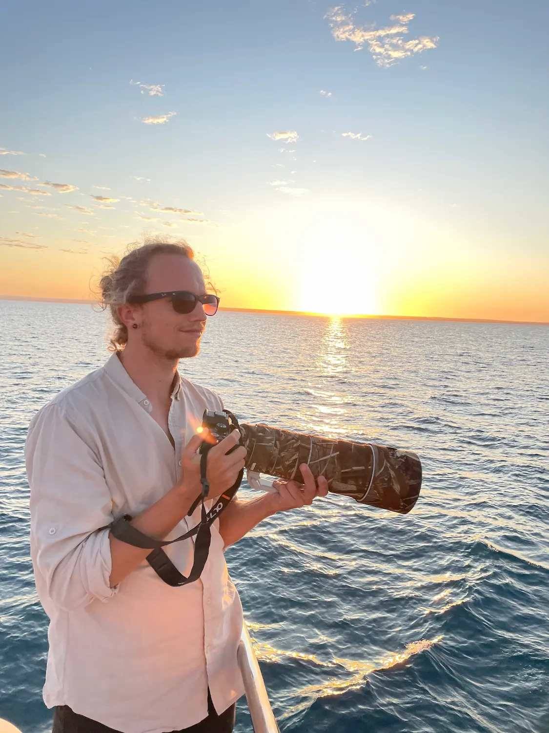 Rainer Lentjes conservation photographer standing on a boat at sunset holding a telephoto camera lens over the ocean.