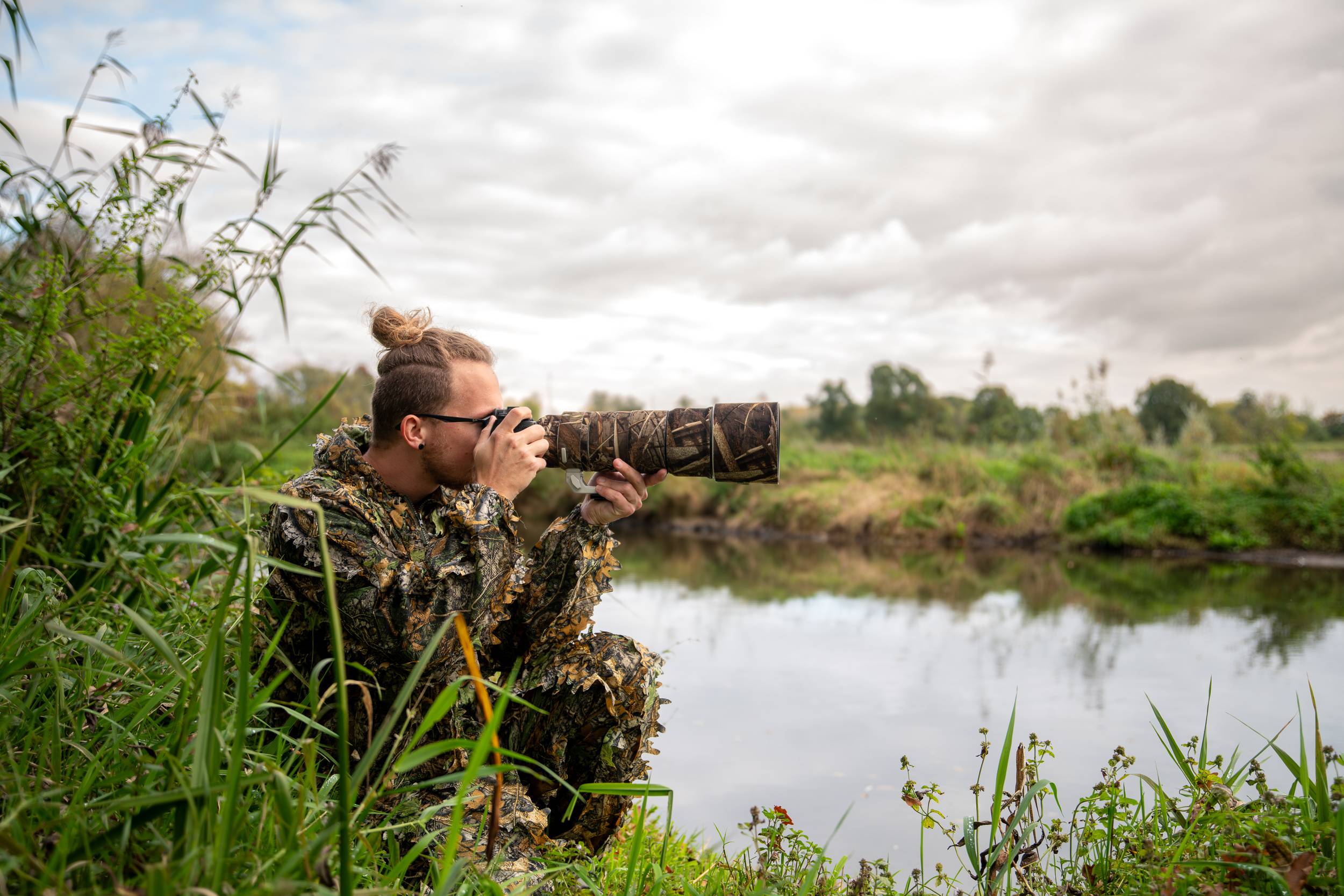 Rainer Lentjes wildlife photographer in camouflage gear crouched near a riverbank capturing nature through a telephoto lens.