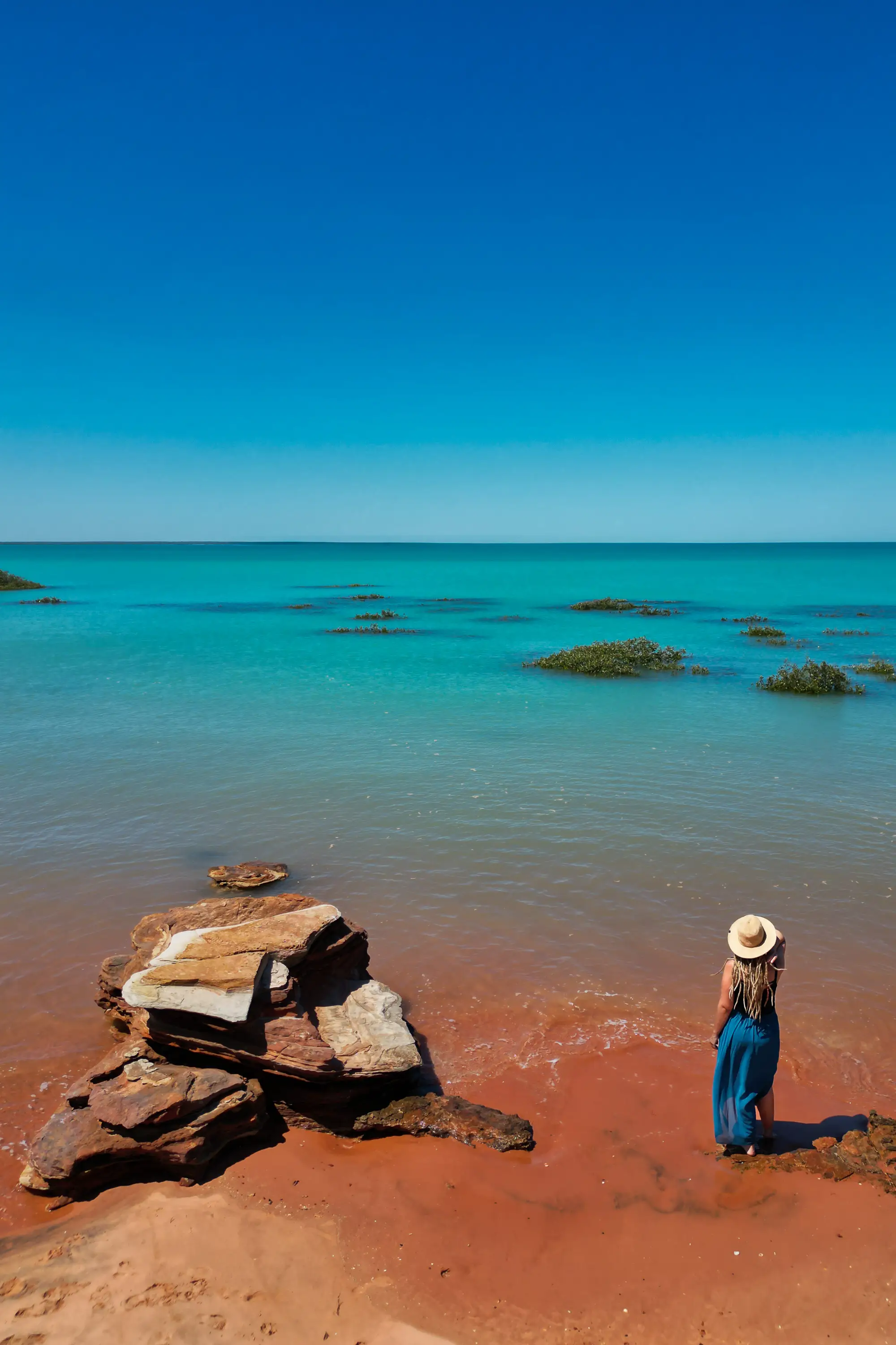 Woman standing on red coastal rocks by turquoise ocean in Broome, Western Australia.